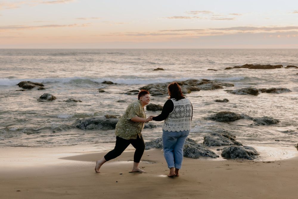 Two people playfully dance together on a rocky beach at sunset with ocean waves in the background.