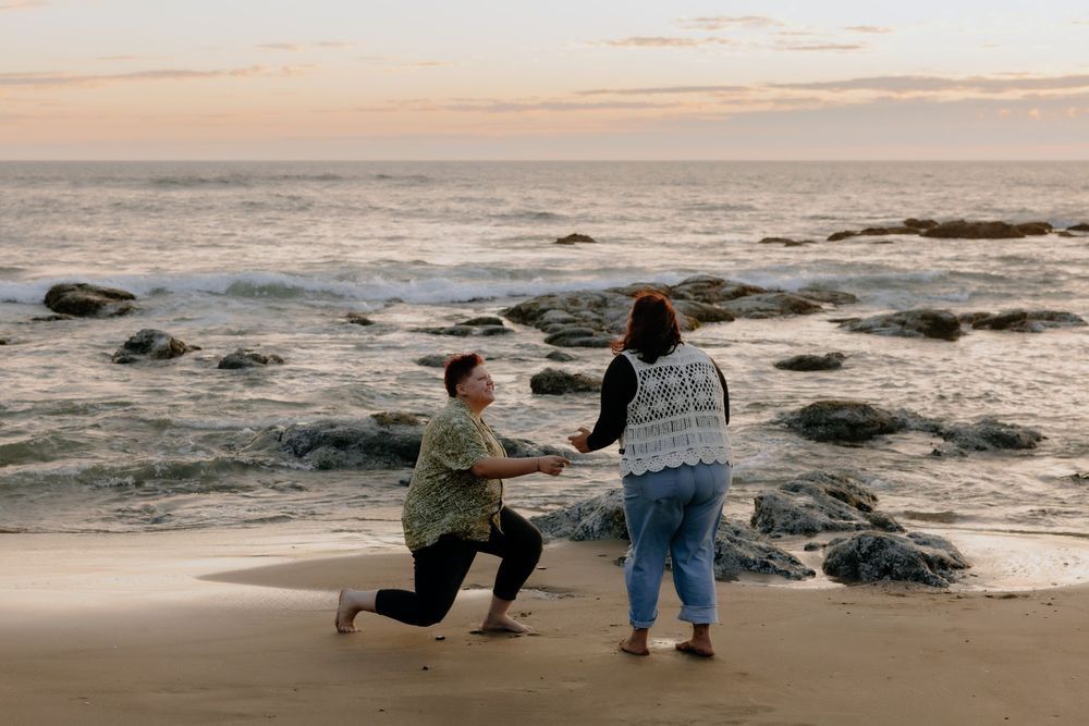 Two people share a romantic moment during sunset on a rocky beach with ocean waves in the background.