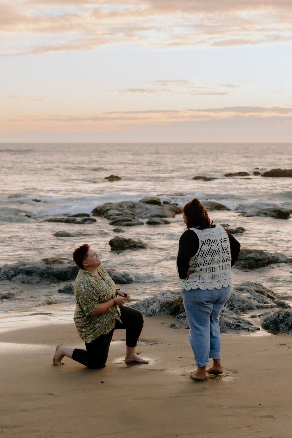 Two people kneel on a beach during golden hour with rocky coastline and ocean waves in the background.