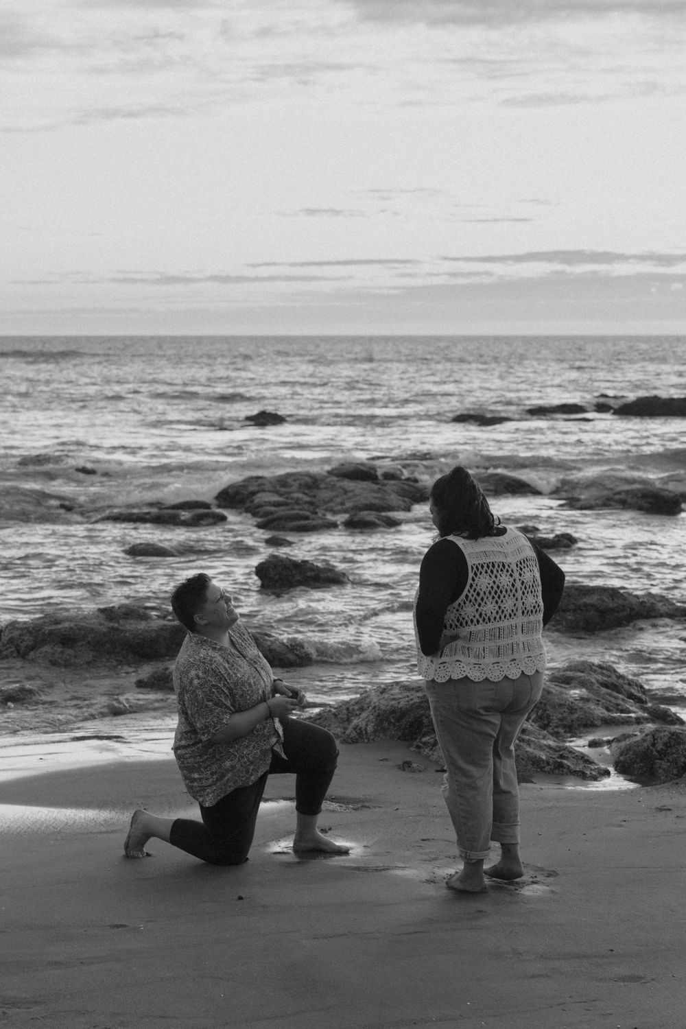 A black and white photograph of two people kneeling on a beach with waves crashing behind them.