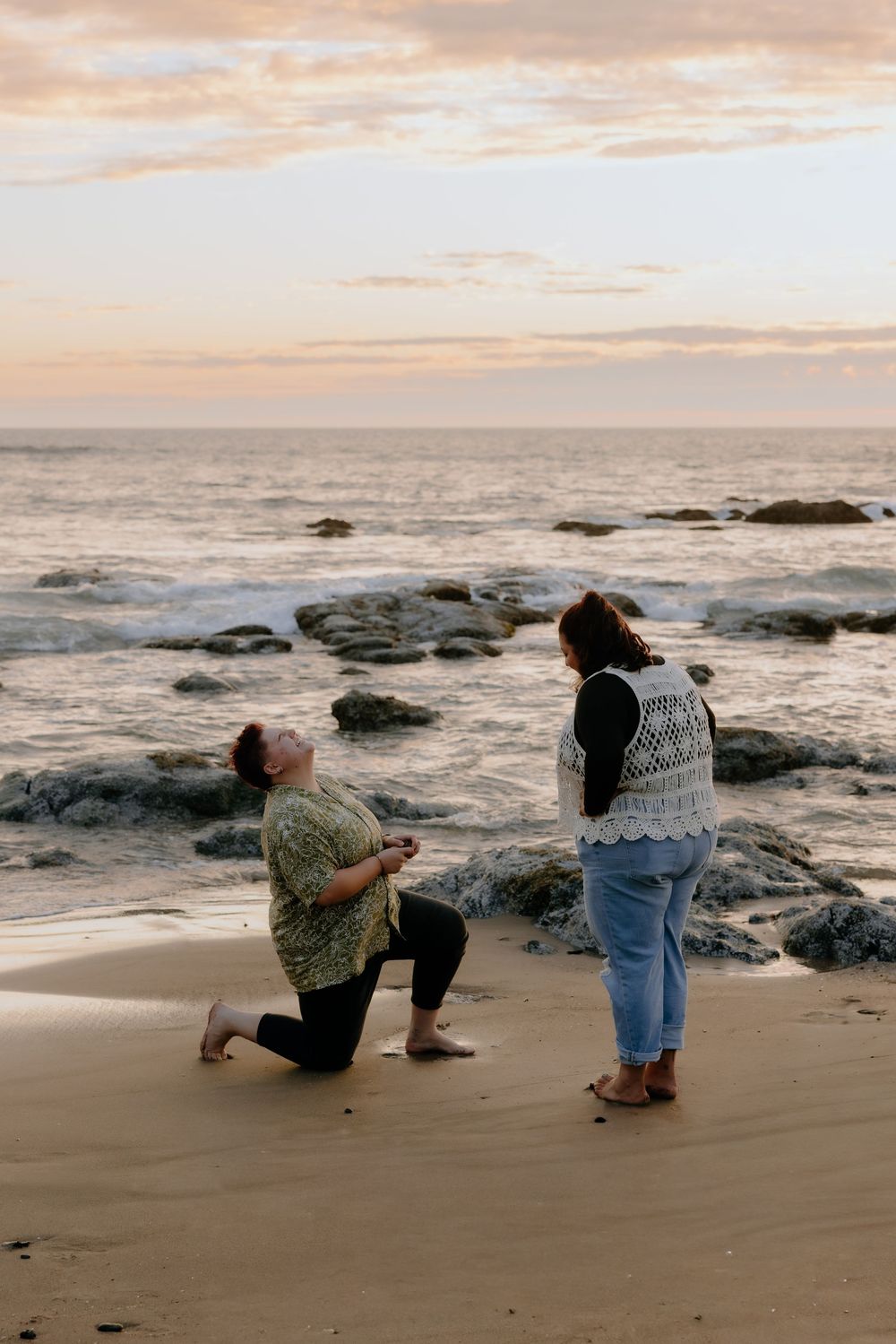 A romantic proposal moment captured at sunset on a rocky beach with crashing waves in the background.