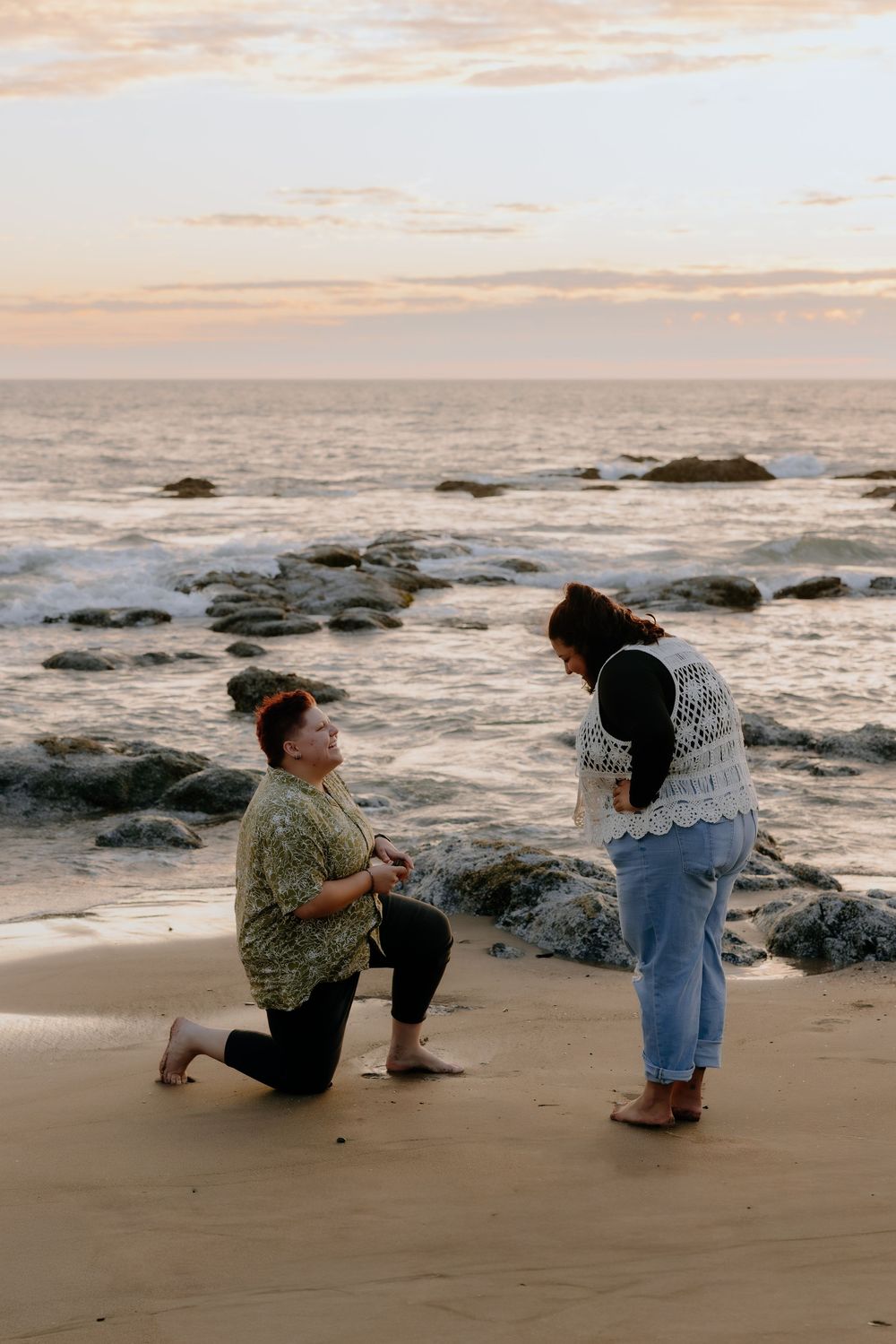 Two figures kneel on a sandy beach at sunset near rocky outcrops and ocean waves.