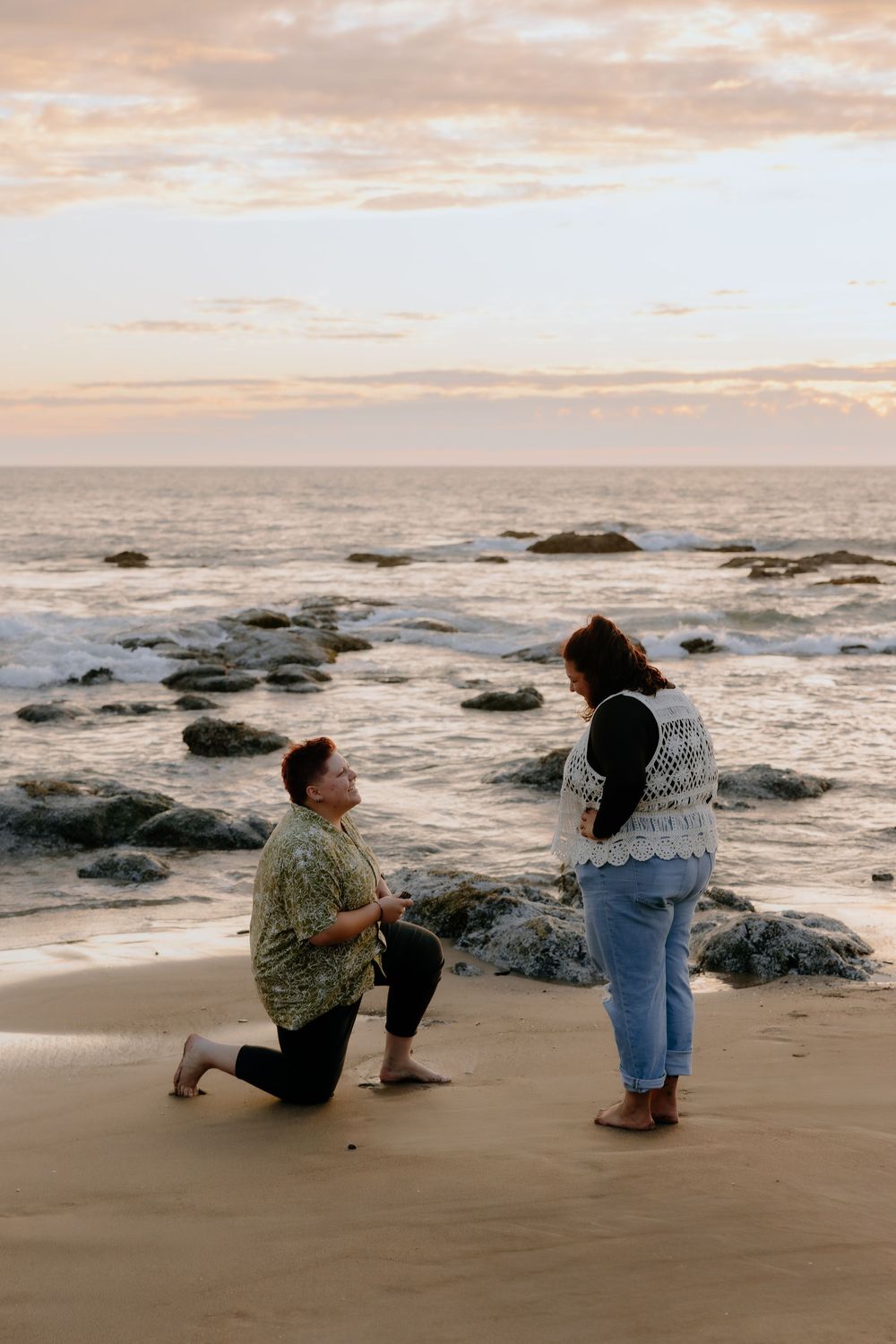 Two people sit on a beach at sunset with waves crashing against coastal rocks in the background.