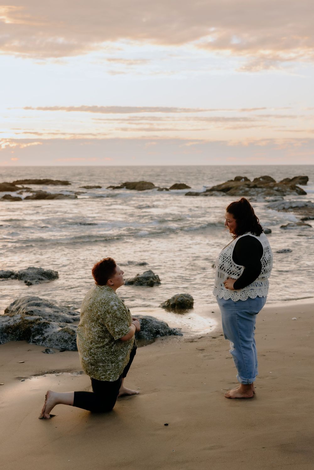 A romantic beach proposal scene with waves and rocks in the background during sunset.