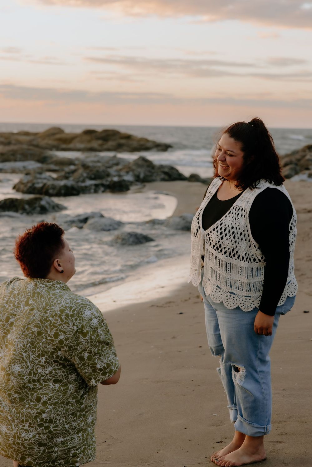 People in casual attire stand on a beach with crashing waves and rocky outcrops at sunset.