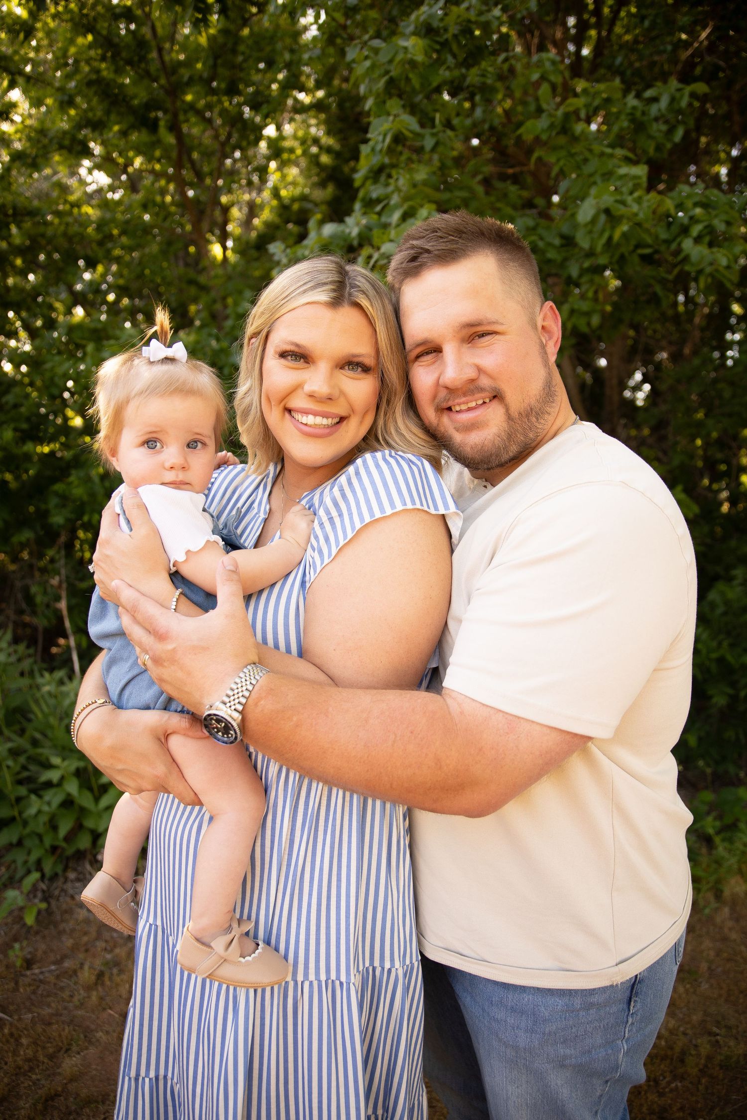 Family wearing coordinated blue and white outfits embraces outdoors during golden hour portrait session.