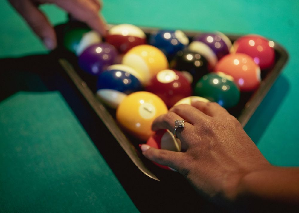 Hands arranging colorful billiard balls in a triangle rack on a green pool table.