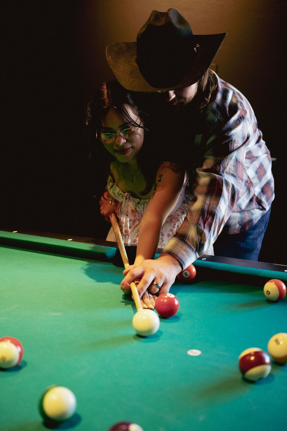 A man in a cowboy hat teaches a woman to play pool, guiding her hands on the cue at a green billiard table.