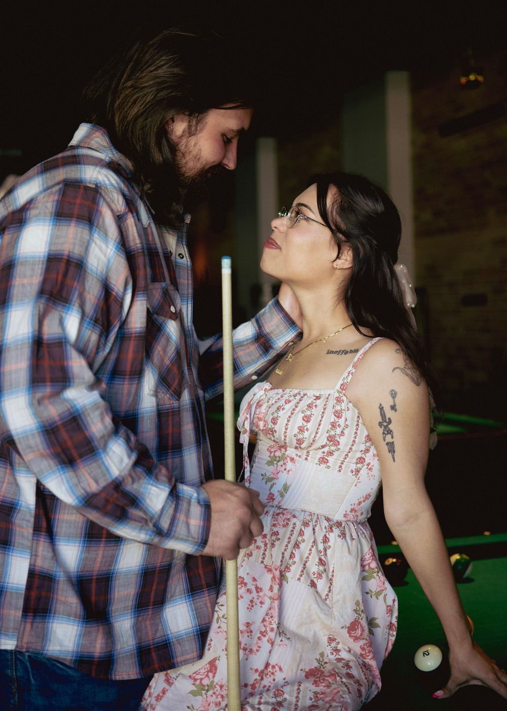 Romantic couple sharing an intimate moment near a pool table, man holding a cue stick in plaid shirt.