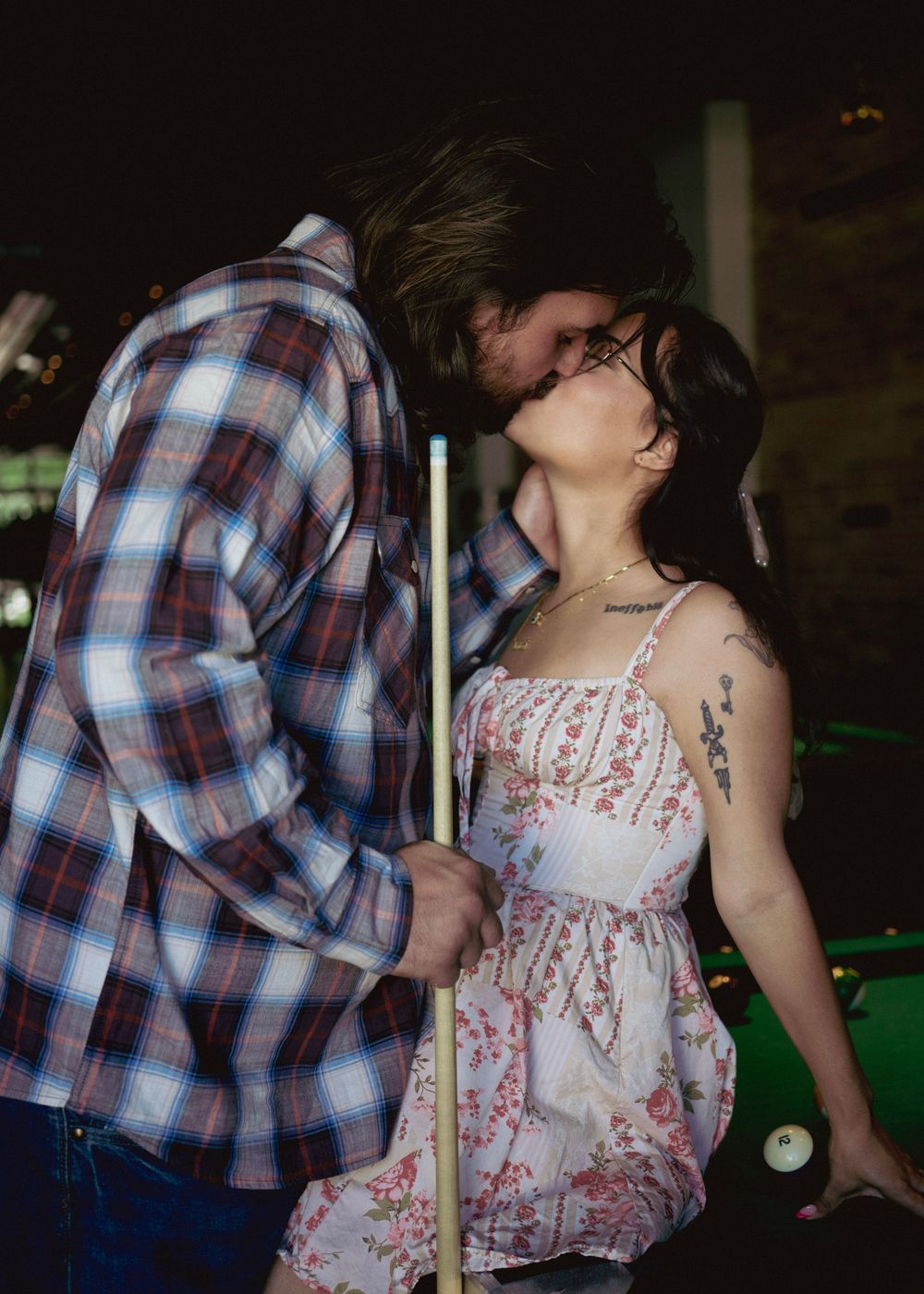 Couple sharing a romantic kiss in a bar, woman in floral dress holding a pool cue, man in plaid shirt.