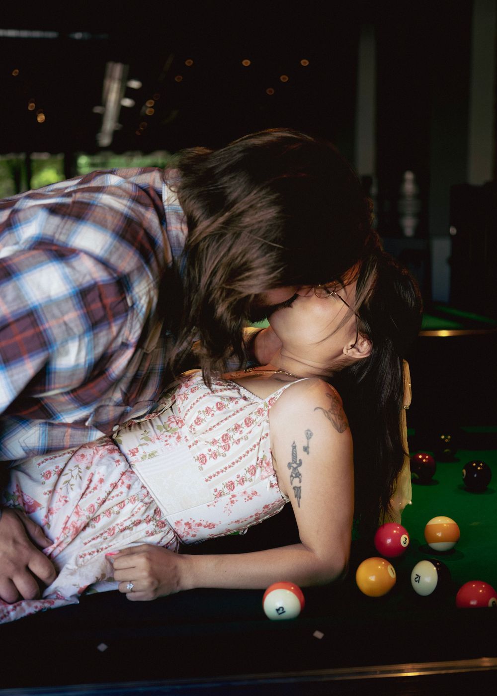 Couple sharing a kiss leaning over a pool table with colorful billiard balls in a bar setting.
