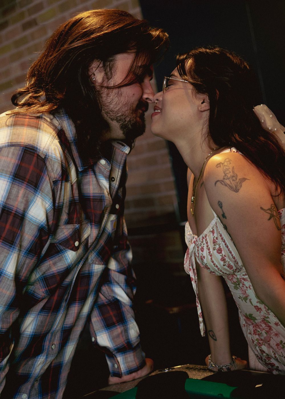 A couple with tattoos sharing a romantic kiss, the man in a plaid shirt and the woman in a floral dress.