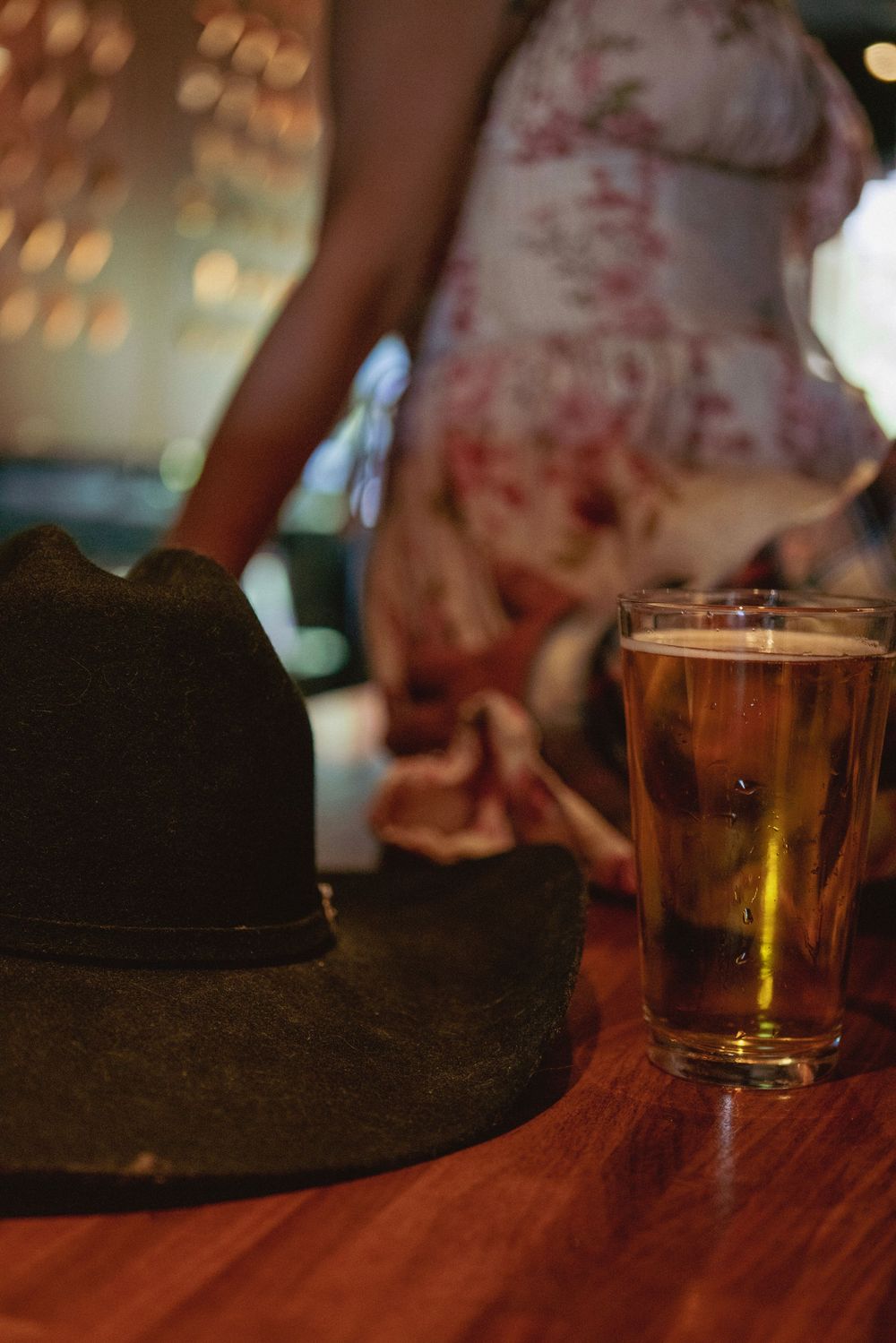 Cowboy hat and glass of beer on wooden bar table with woman in floral dress blurred in background.