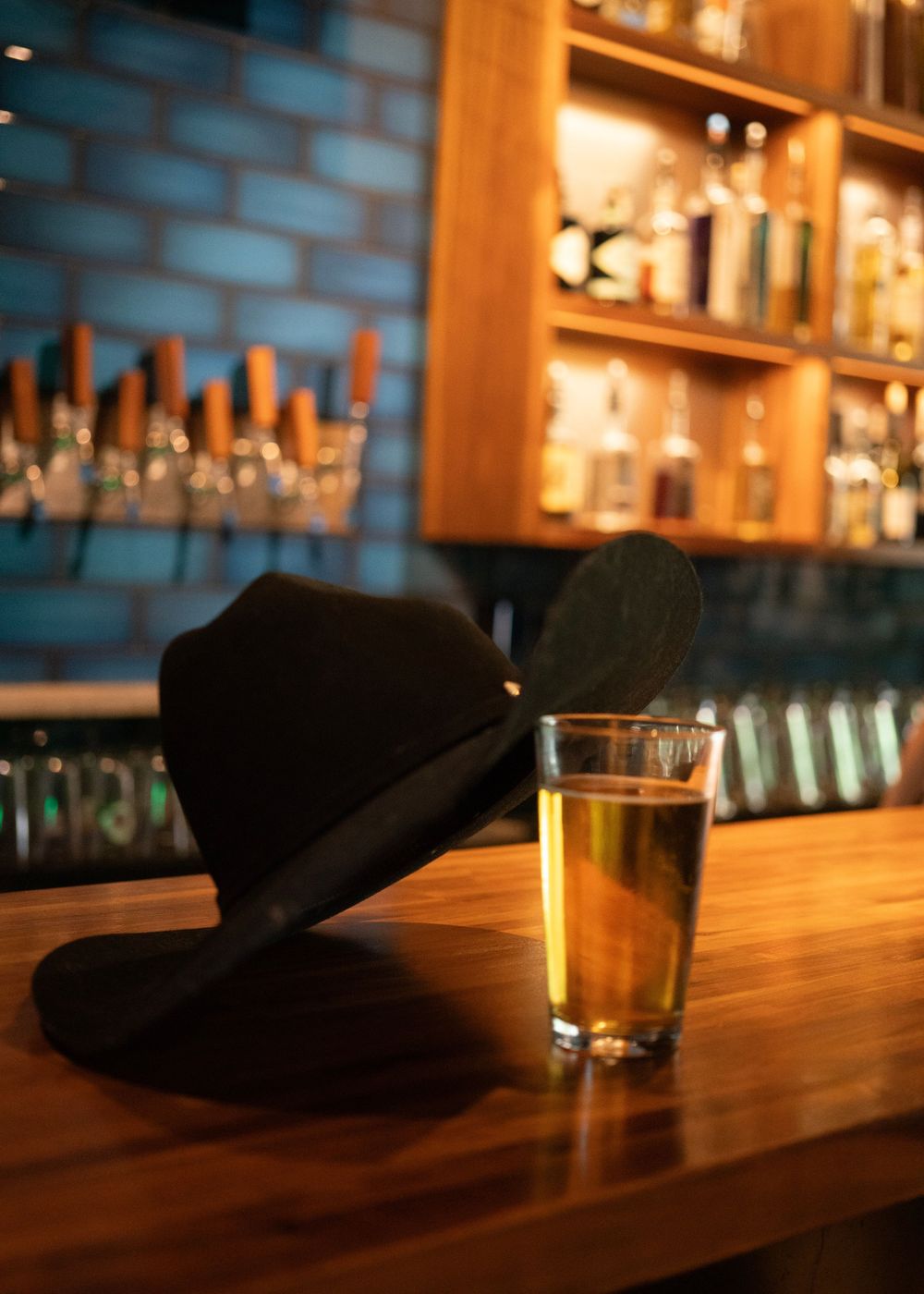 A black fedora hat rests beside a glass of beer on a wooden bar counter with liquor shelves in background.