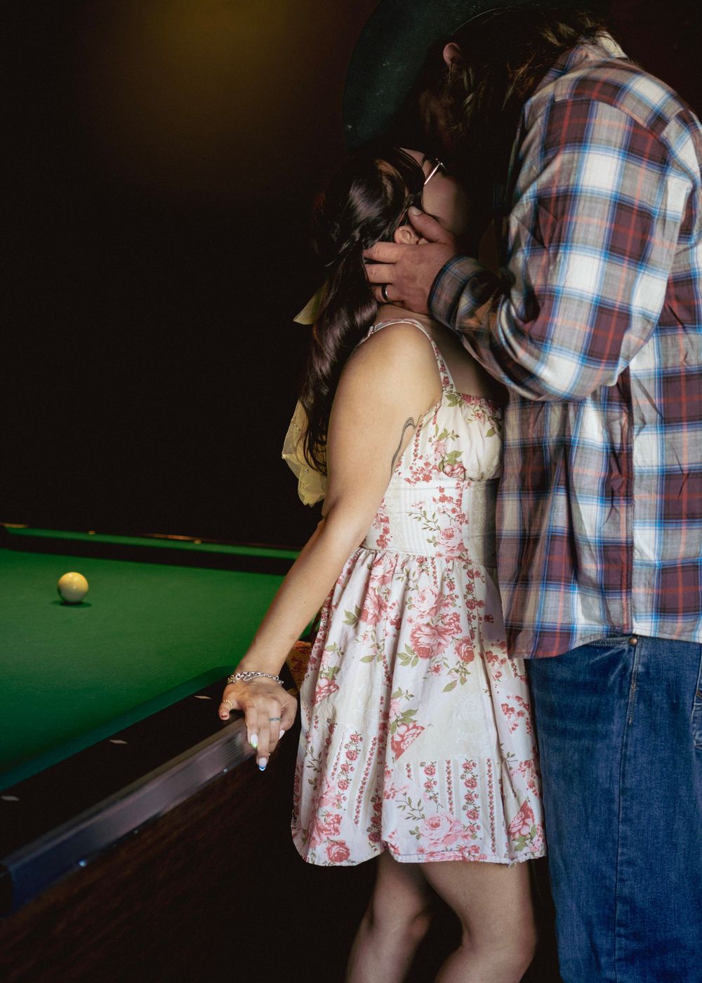 Couple kissing in floral dress and plaid shirt by a green pool table in a dimly lit bar.