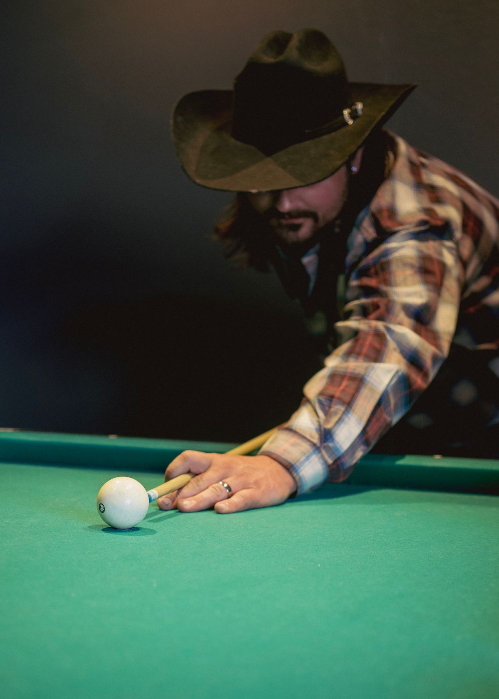 Man in cowboy hat and plaid shirt playing pool, leaning over green billiard table to shoot cue ball.