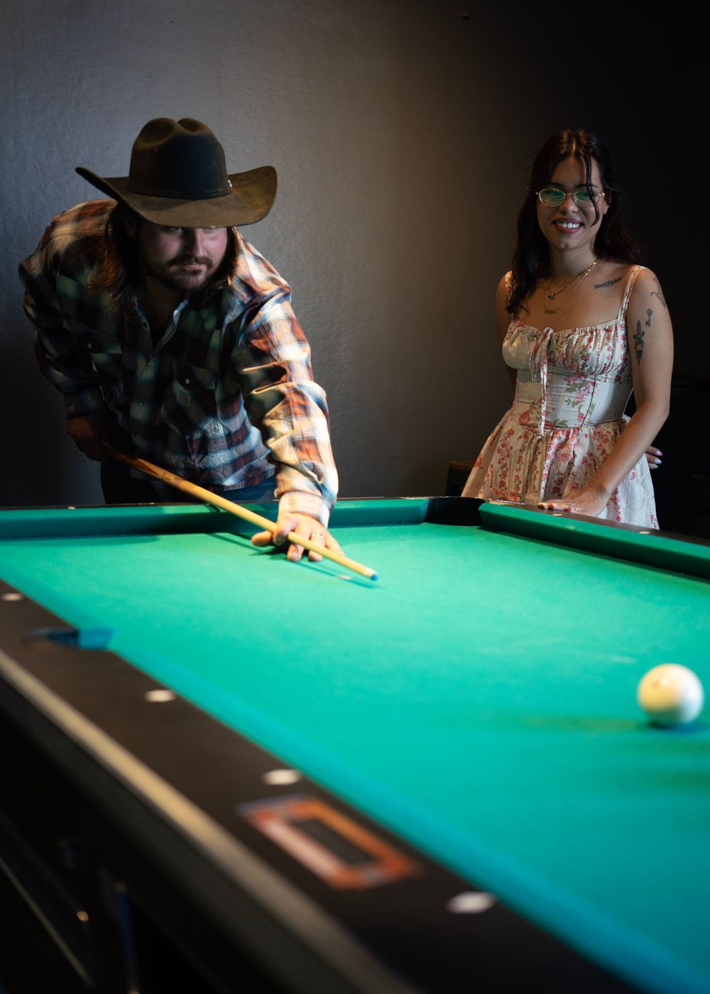 Man in cowboy hat playing pool while woman in floral dress watches and smiles in a dimly lit bar.