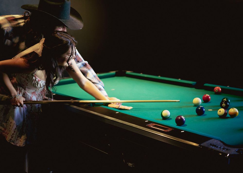 Woman in floral dress and hat leaning over green pool table taking a shot surrounded by billiard balls.