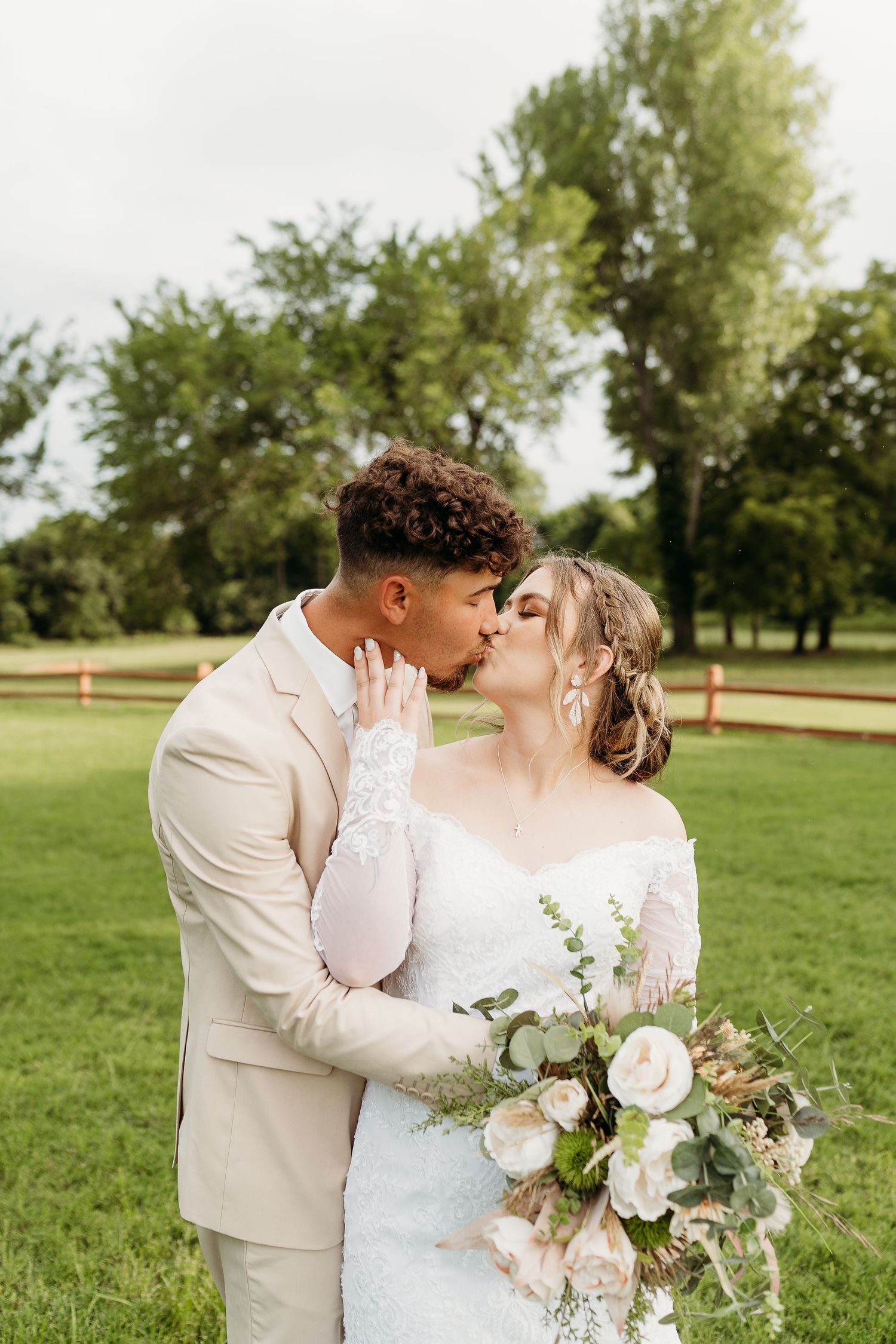 A bride and groom share an intimate kiss outdoors on their wedding day while holding a bouquet of white roses.