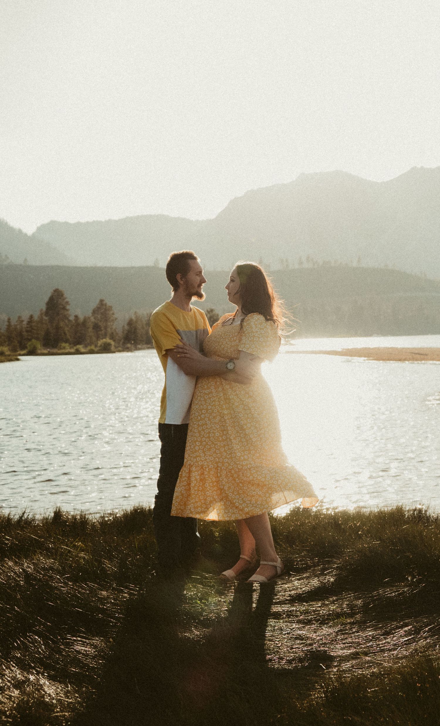 A couple shares a romantic sunset moment by a mountain lake with golden sunlight streaming through the scene.