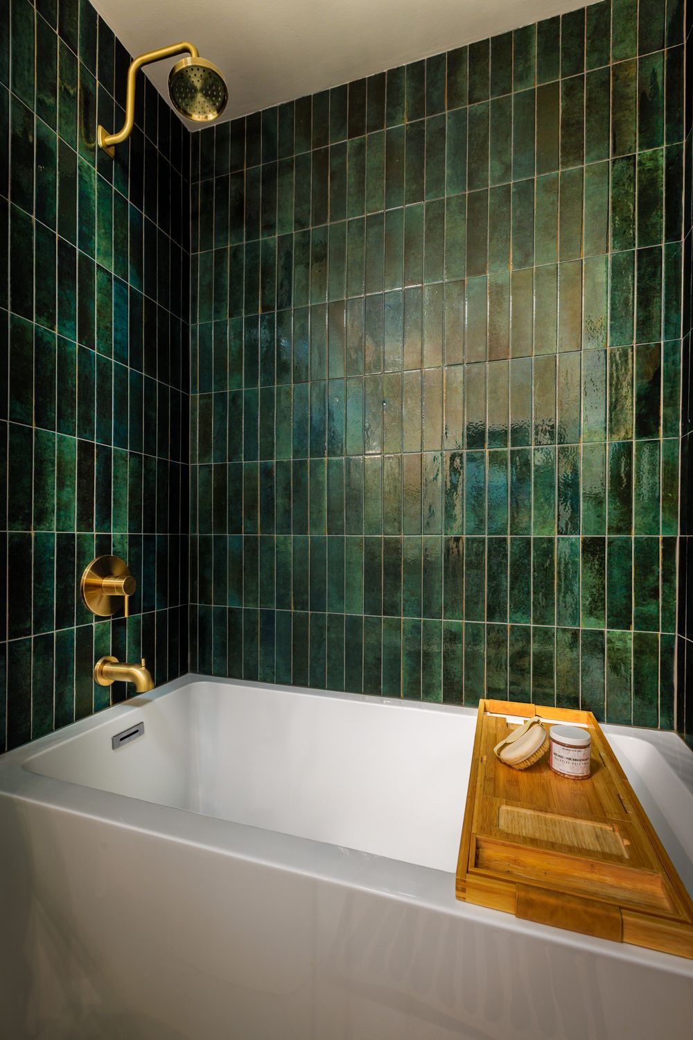 White bathtub with brass fixtures surrounded by lush green zellige tiles and a wooden bath caddy with accessories.