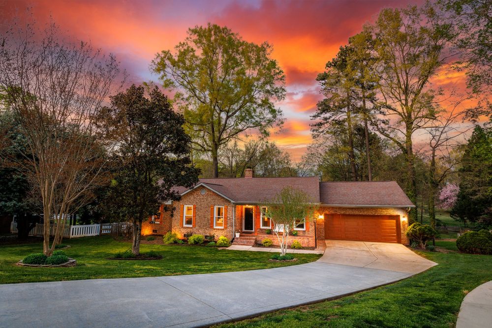 Ranch home with brick exterior, attached garage, lush green lawn, and vibrant sunset sky surrounded by tall trees.
