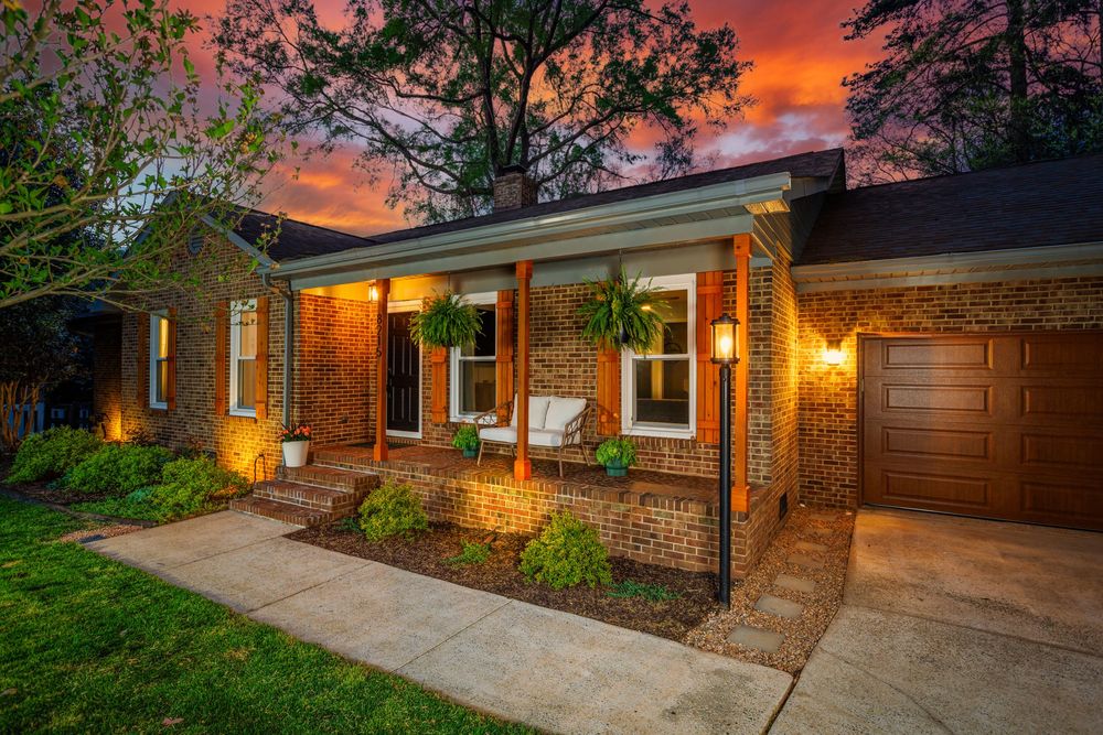 Charming brick ranch home with covered porch, lush landscaping, and warm exterior lighting under a vibrant sunset sky.