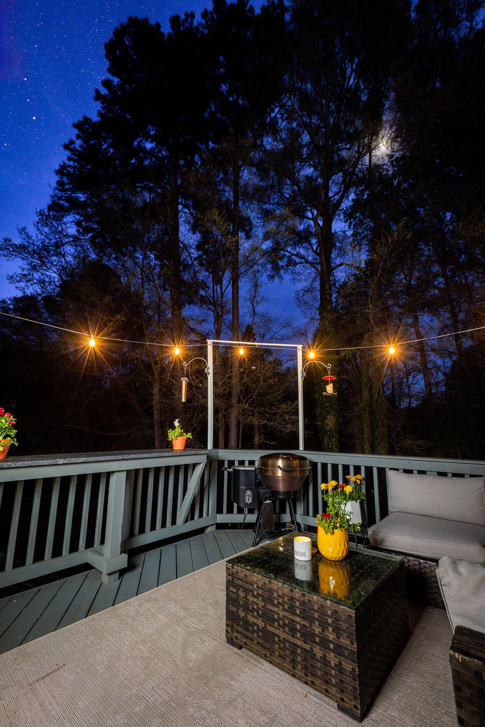 Outdoor deck at night with string lights, wicker furniture, and a starry sky surrounded by tall trees.