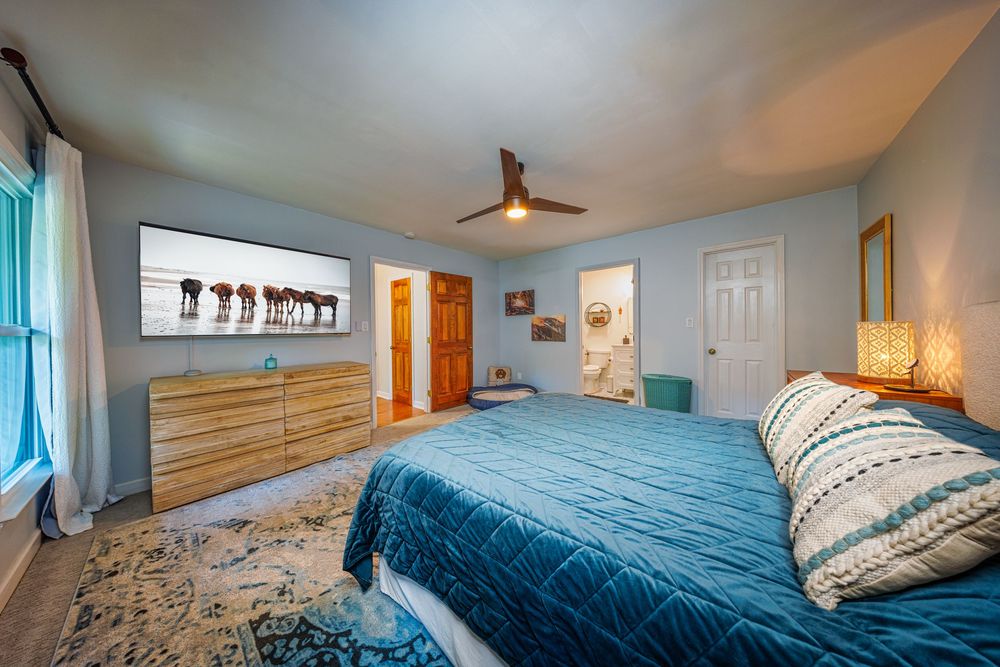 Cozy bedroom with teal bedding, wooden dresser, ceiling fan, and horse artwork above a large window.