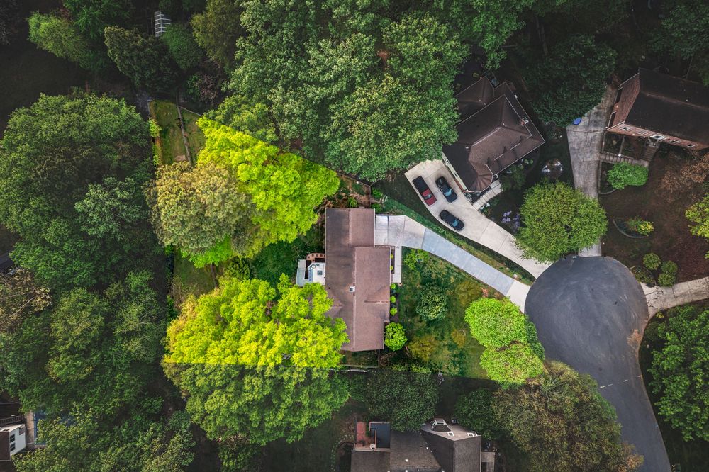 Aerial drone view of suburban homes surrounded by lush green trees with a winding driveway and parked cars visible.