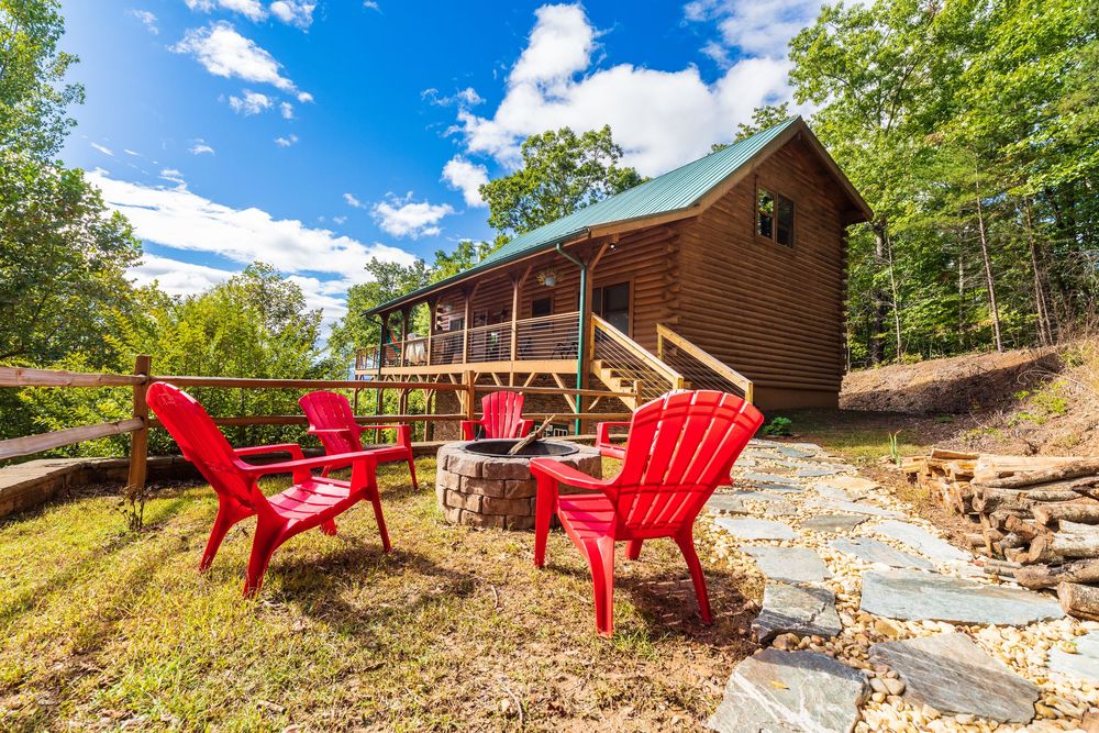 Log cabin with red Adirondack chairs on the front porch and stone pathway leading to entrance.