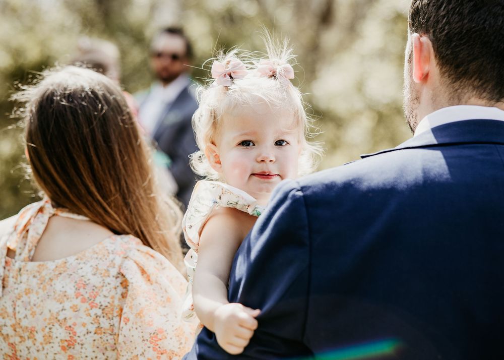Adorable baby girl in floral dress with pink bow is held by parents during an outdoor wedding ceremony.