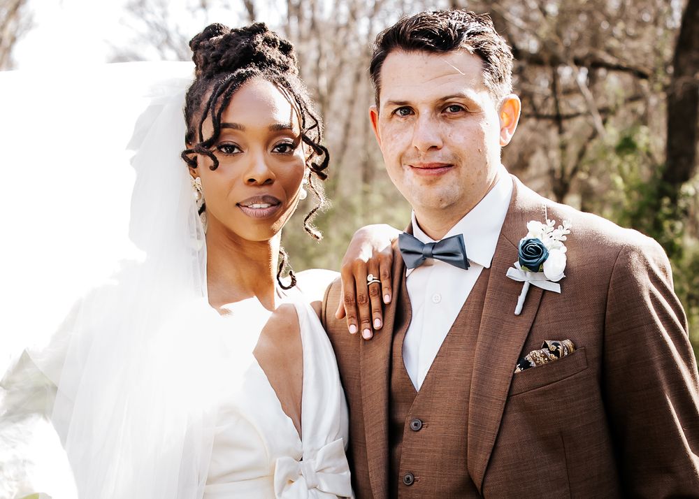 Interracial wedding couple smiling together, bride in white veil and groom in brown suit with blue bowtie.