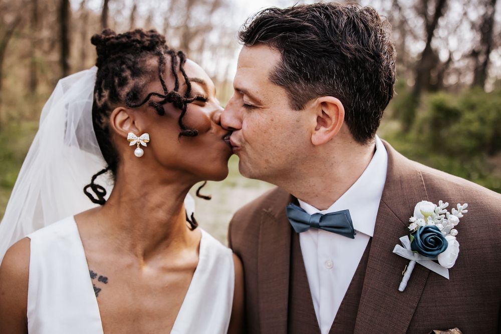 Interracial bride and groom sharing a romantic kiss on their wedding day, groom wearing brown suit with blue boutonniere.