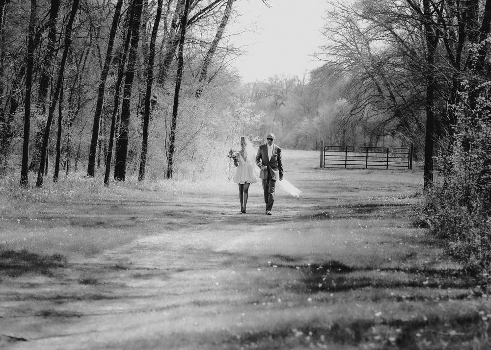 Black and white photo of bride and groom walking down a tree-lined path.