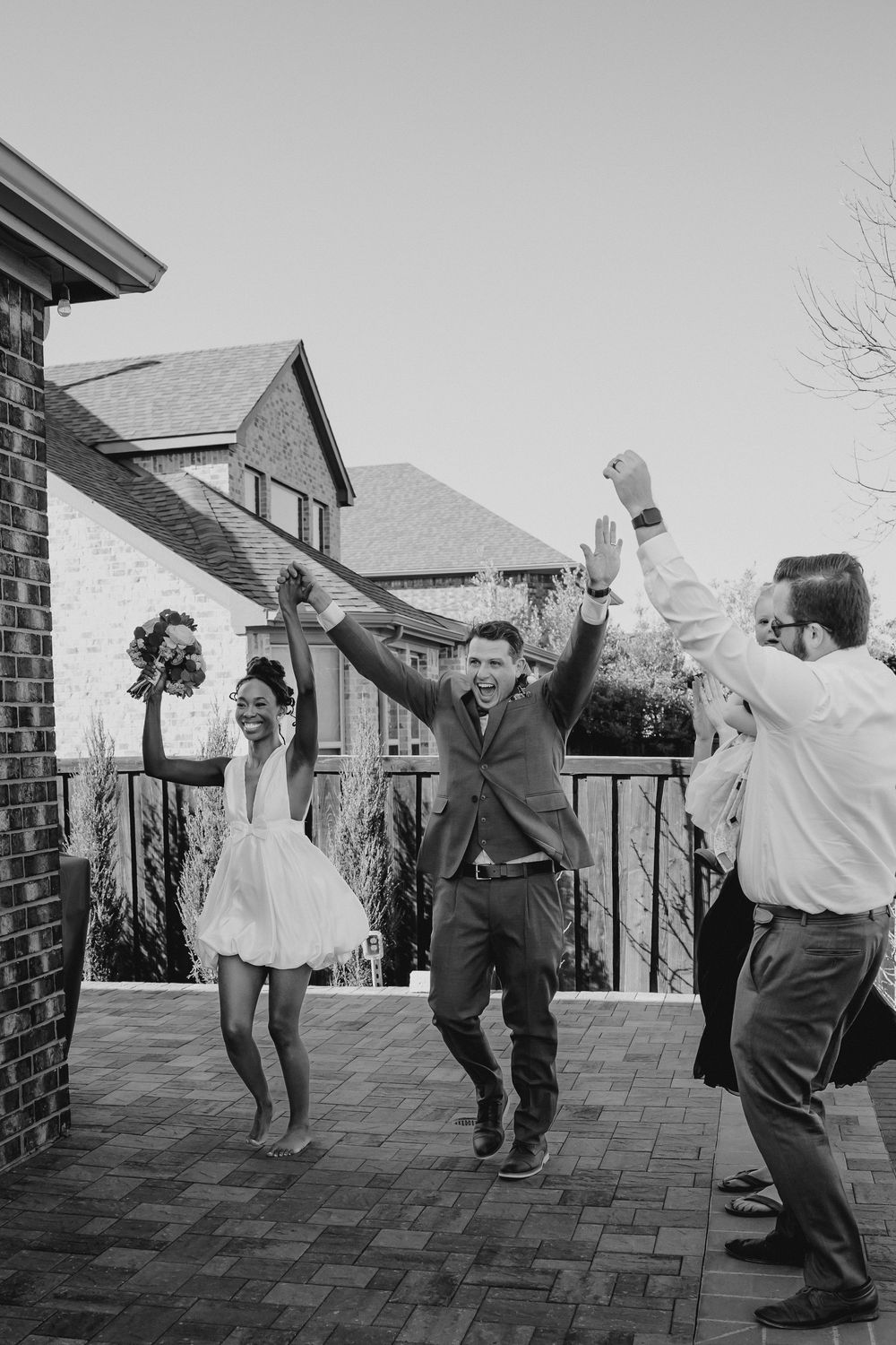 Black and white photo of newlyweds celebrating with raised hands outside with wedding guests.