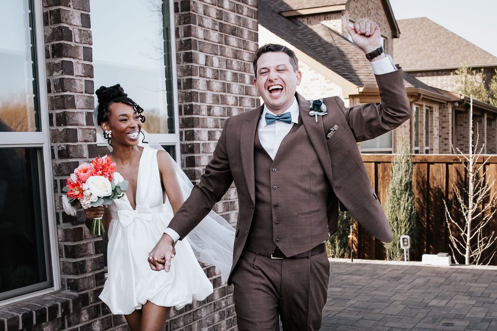 Joyful newlywed couple celebrating outside brick building, bride holding colorful bouquet, groom raising fist in excitement.