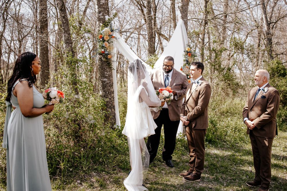 Wedding party gathers at floral arch for outdoor woodland ceremony with officiant.