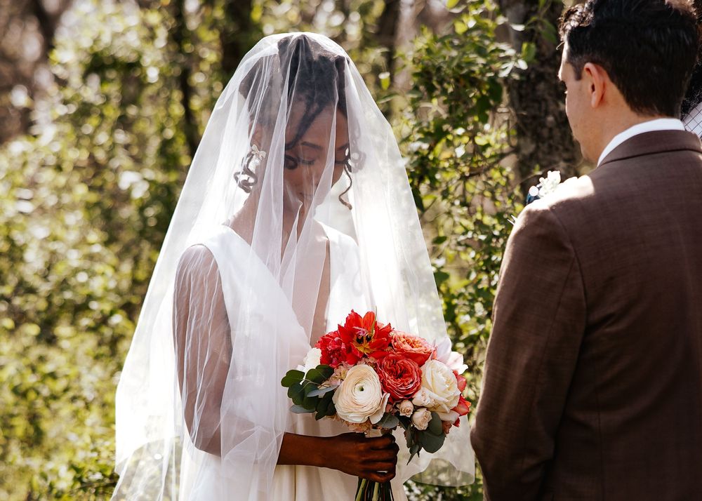 Veiled bride in white gown holds colorful bouquet facing groom in brown suit.