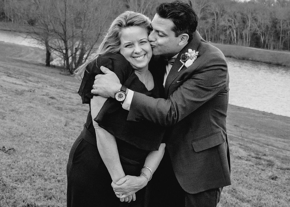 Black and white photo of a groom kissing bride on cheek while embracing near a lake.