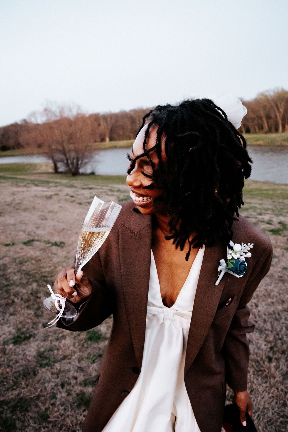 Joyful bride with locs in white dress and brown blazer holding champagne flute outdoors.