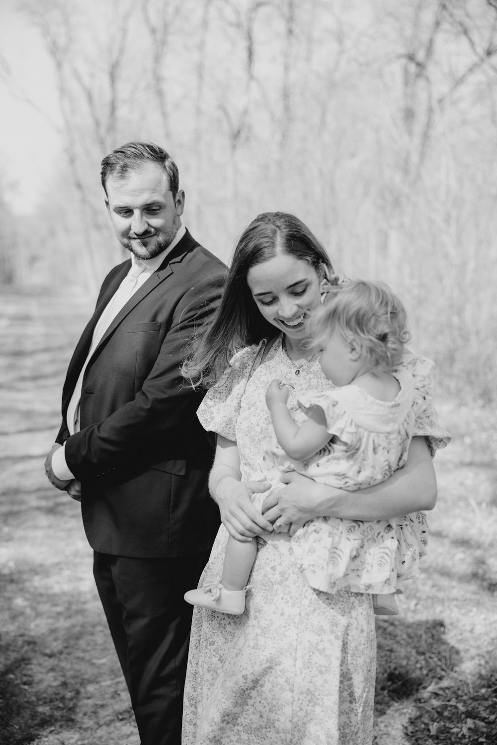 Black and white photo of smiling couple holding baby in wooded outdoor setting.