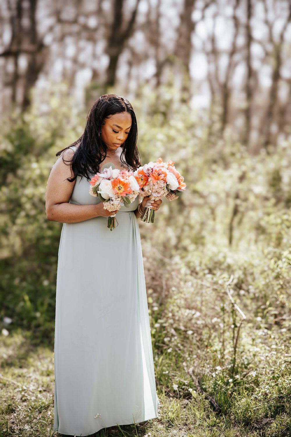 Bridesmaid in mint gown holds coral and white bouquets in a sunlit outdoor garden setting.