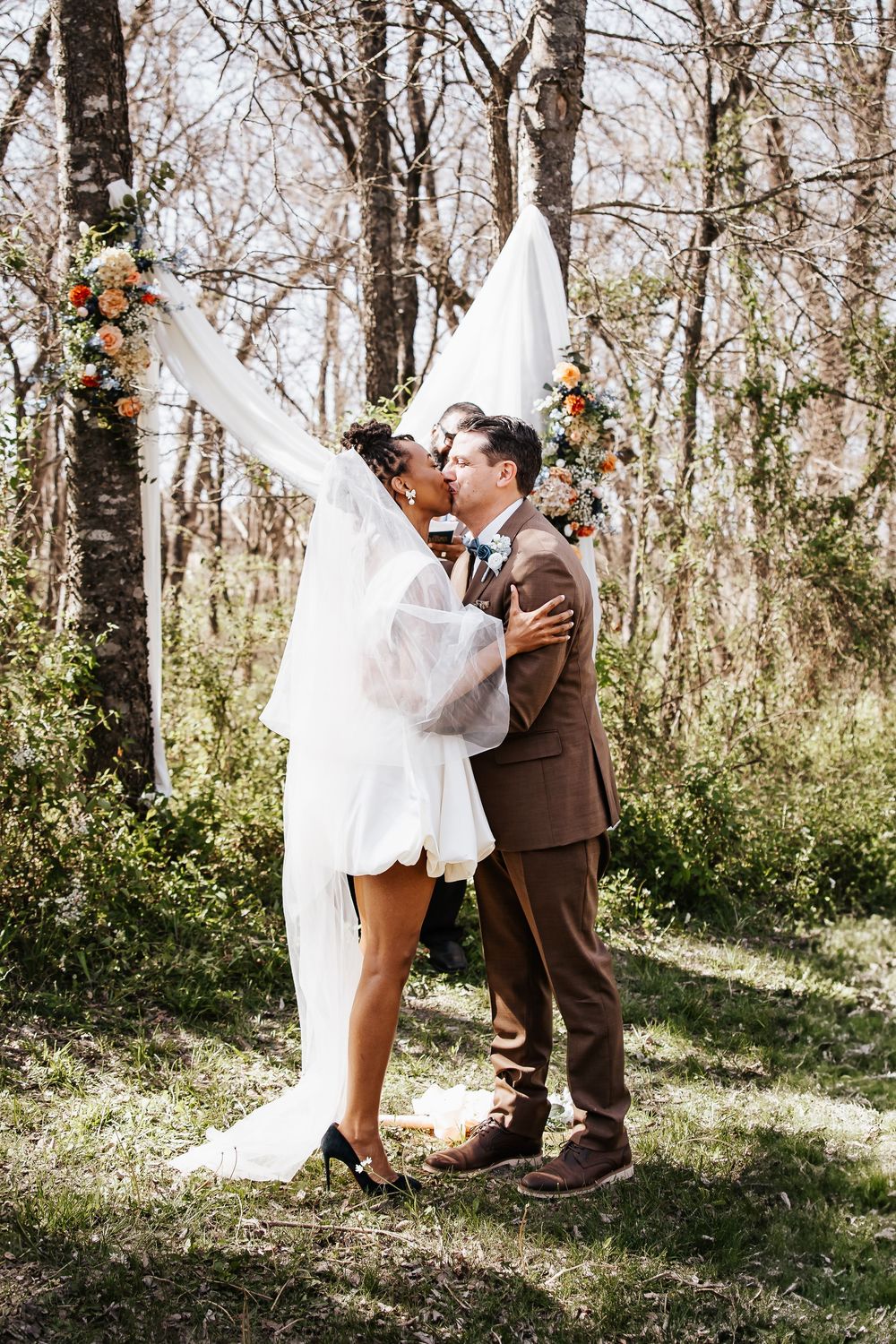 Newlywed couple kisses under flowing white drape at outdoor woodland wedding ceremony.