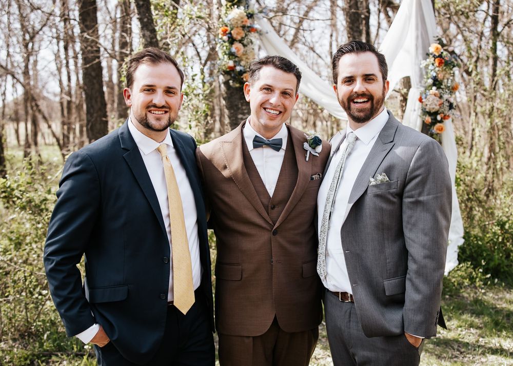 Groom poses with two groomsmen in suits in front of floral wedding arch outdoors.