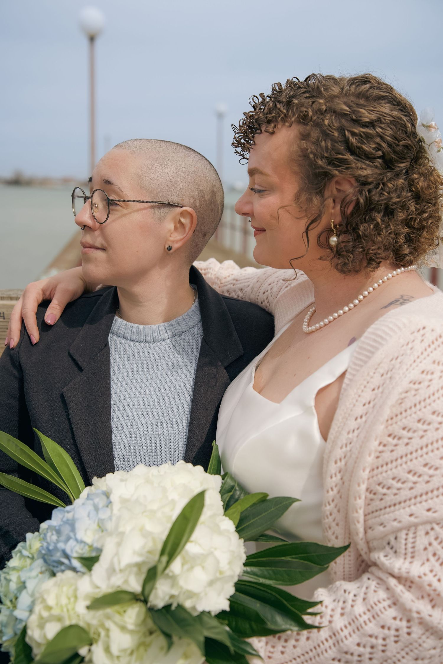 A couple shares an intimate moment outdoors, with white hydrangea bouquet and one wearing glasses and a gray sweater.