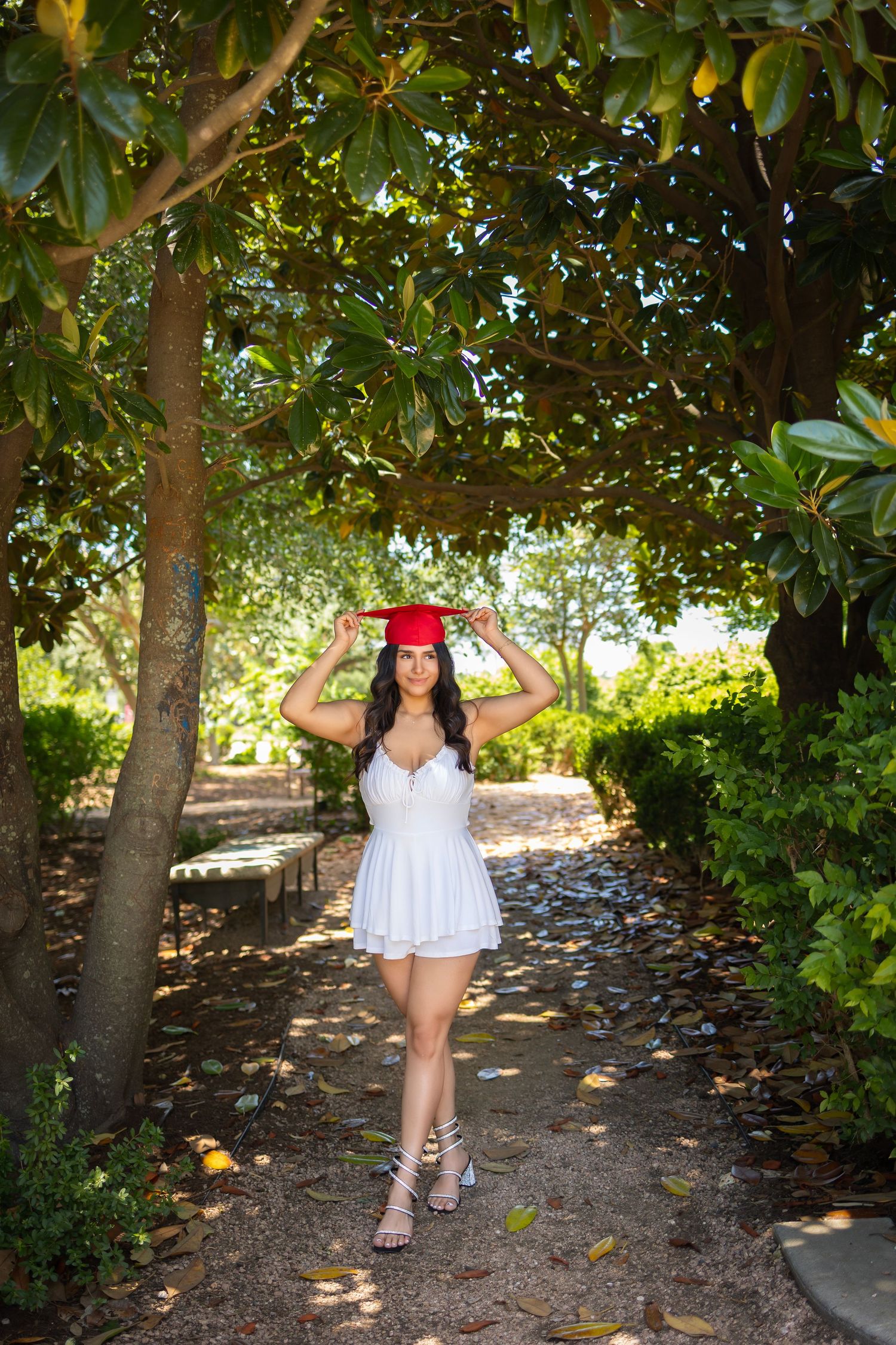 A figure in a white summer dress and red hat poses on a tree-lined garden path on a sunny day.