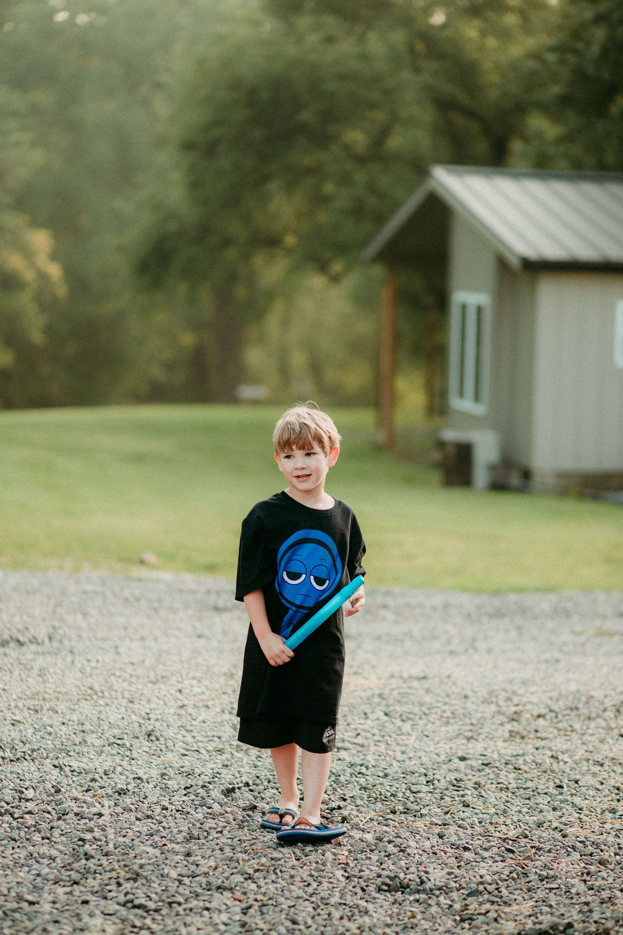 A child in a black t-shirt with a blue design stands on a gravel path near a small shed on a summer day.