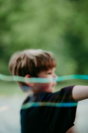 A child in a black t-shirt stands outdoors shooting a toy bow and arrow against a blurred green background.
