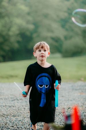 Child in black t-shirt with blue cartoon design plays with bubbles on gravel path surrounded by green trees.